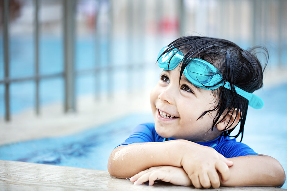 Enfant souriant de 4-6 ans dans l'eau d'une piscine municipale, accoudé sur le bord du bassin
