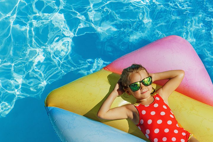 Petite fille avec lunettes de soleil et maillot de bain rouge à pois faisant la sieste sur un matelas gonflable qui flotte dans une piscine.